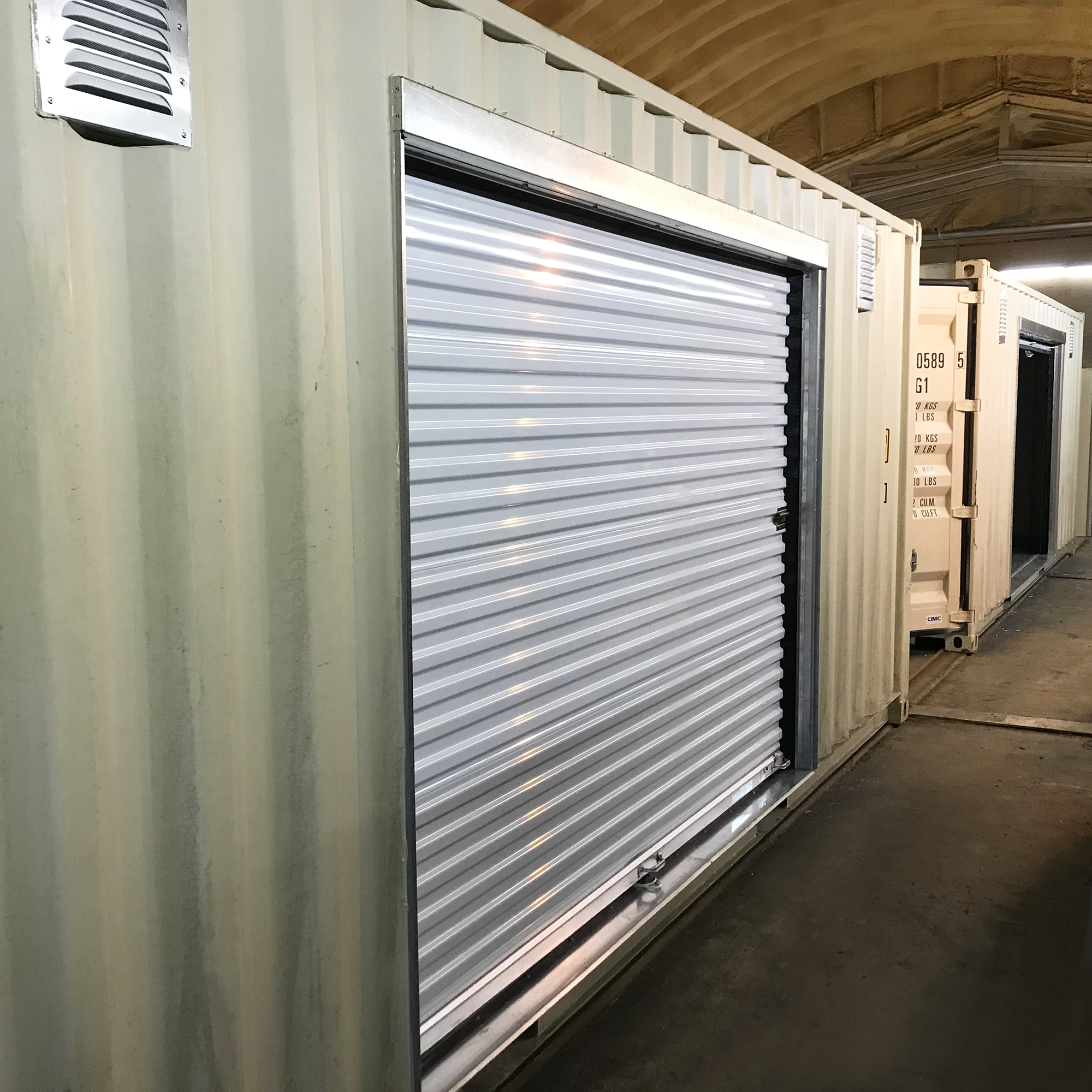 Angled view of a white roll-up Janus 650 door installed on the side of a beige shipping container using a galvanized steel framing kit. Ideal for container conversions and storage units; only the framing kit is sold separately.