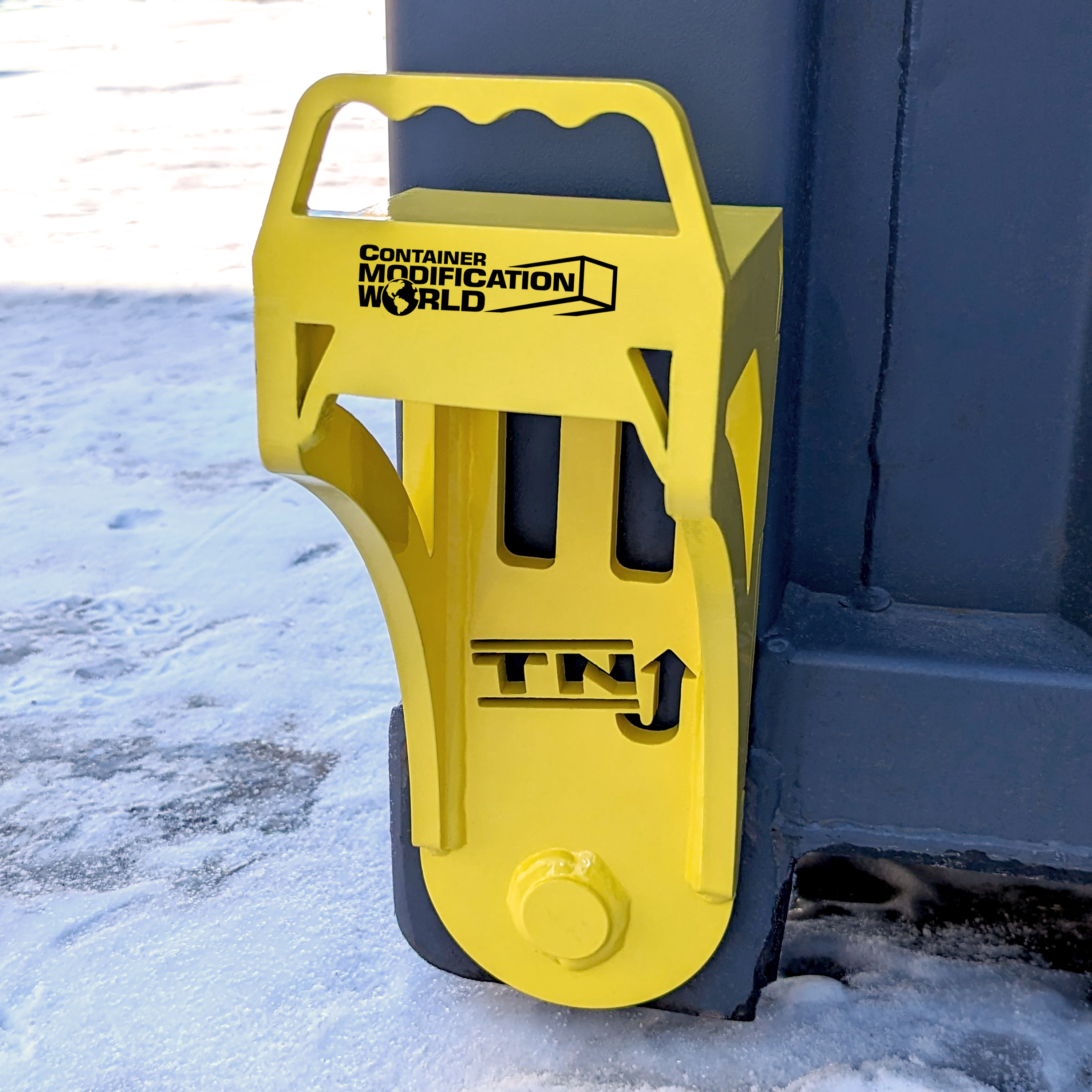 Bright yellow steel container jacker labeled "Container Modification World" and "TNJ," shown in use on a shipping container corner to lift or level it on a snowy surface.