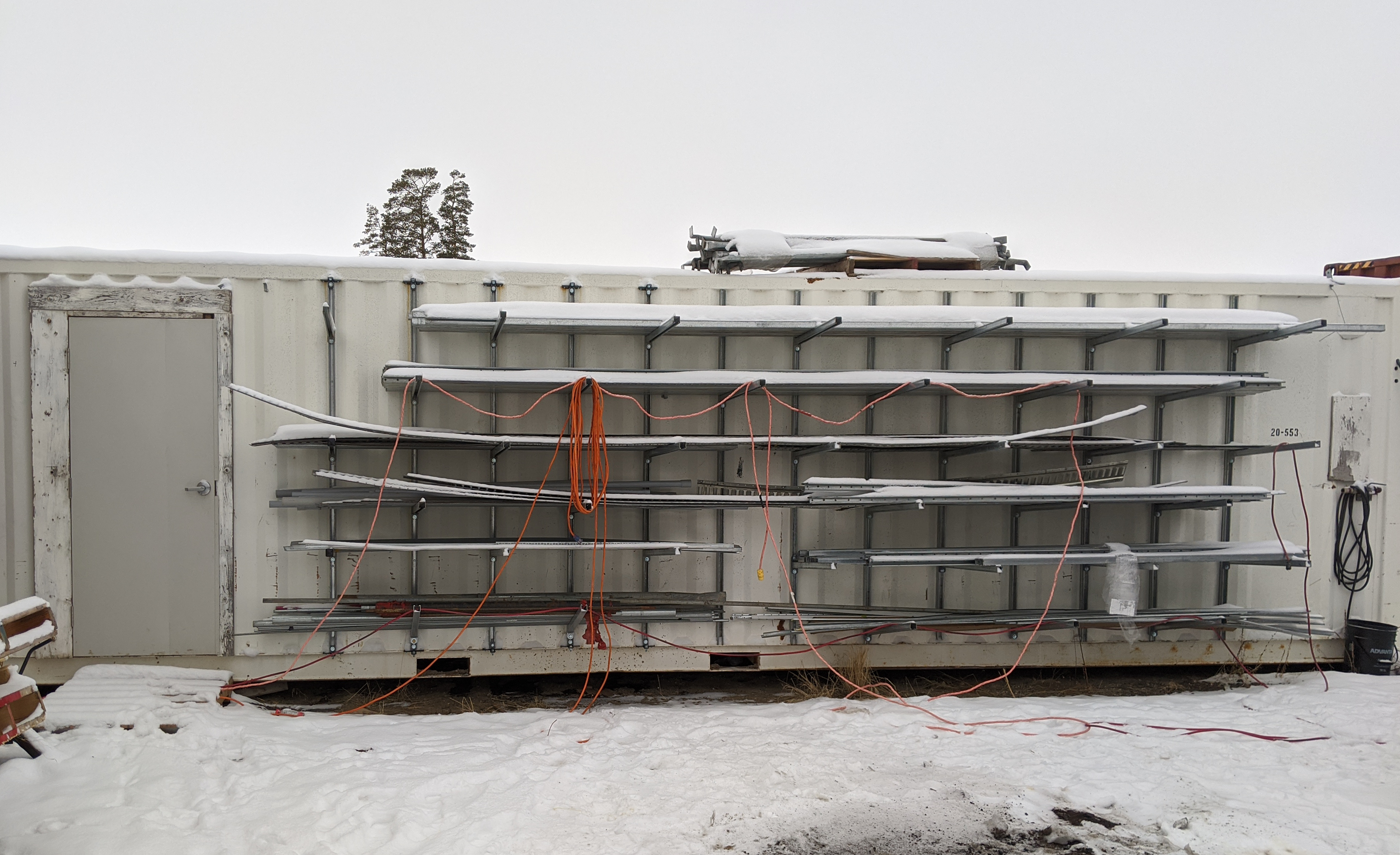 Outdoor side view of a shipping container with heavy-duty galvanized pipe rack brackets and strut system installed, holding multiple layers of long metal conduit in snowy conditions.