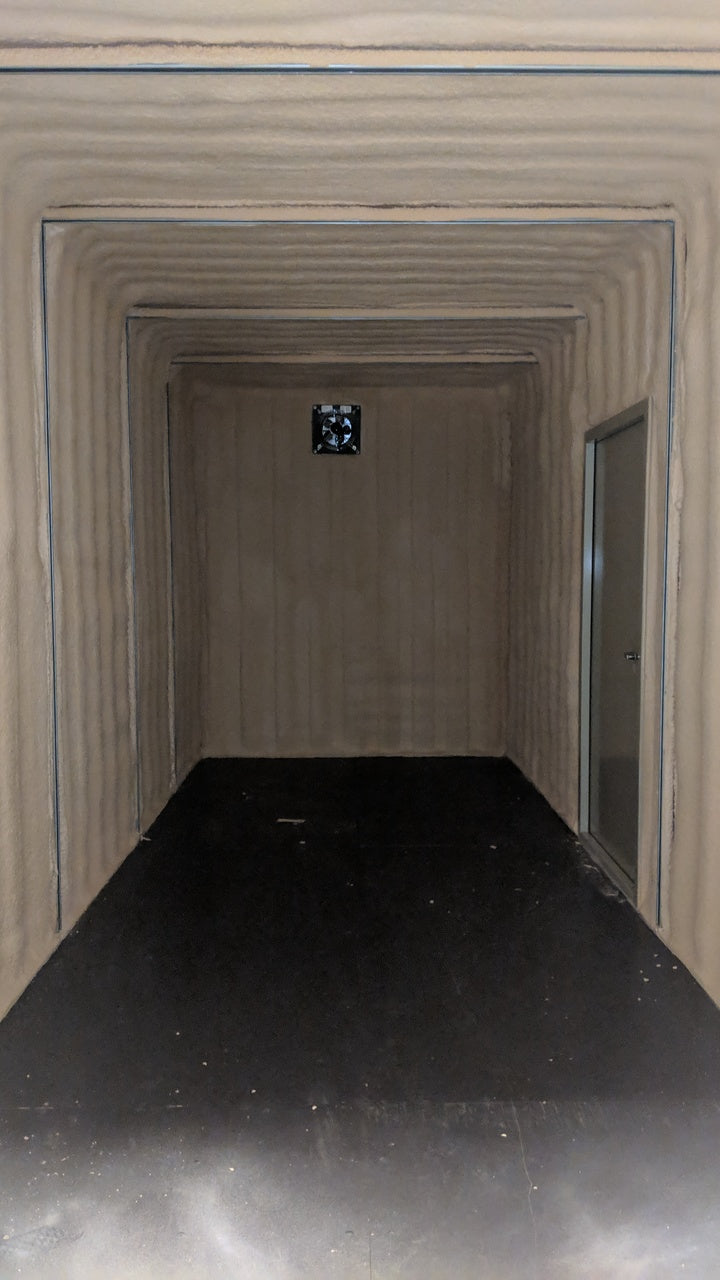 Interior of an insulated shipping container featuring spray foam insulation, a ventilation fan on the back wall, and a framed man door on the right side.