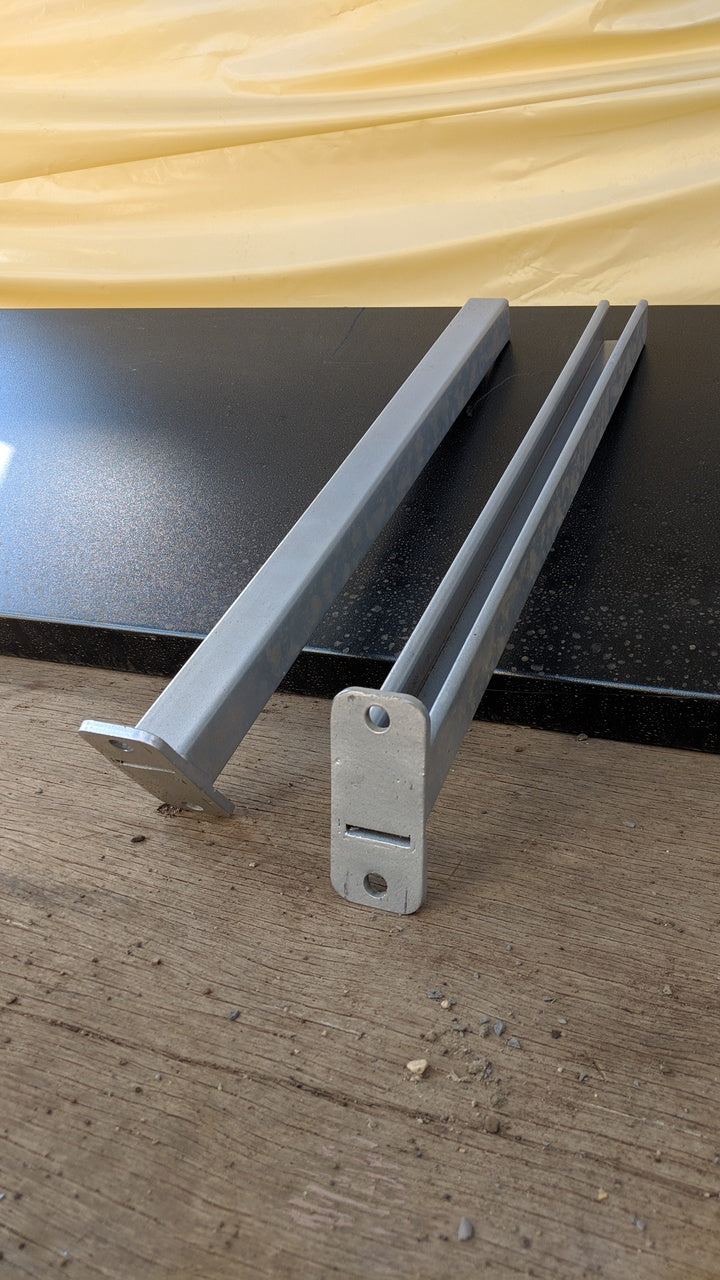 Two galvanized CSM pipe rack brackets for shipping container strut systems, displayed on a black surface with wood flooring — front and angled views showing slotted mounting plates.